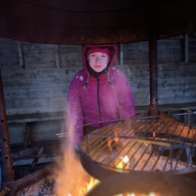 Girl in a pink winter jacket smiling behind a burning campfire grill inside a wooden hut shelter.