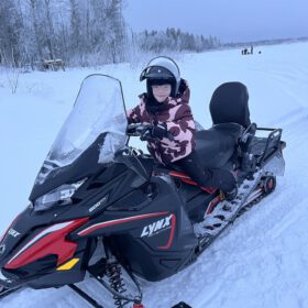 Young child wearing a helmet sitting on a black Lynx snowmobile on a snowy winter trail.