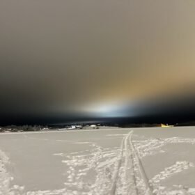 Snow tracks leading across a frozen lake at night toward glowing lights under an overcast winter sky.