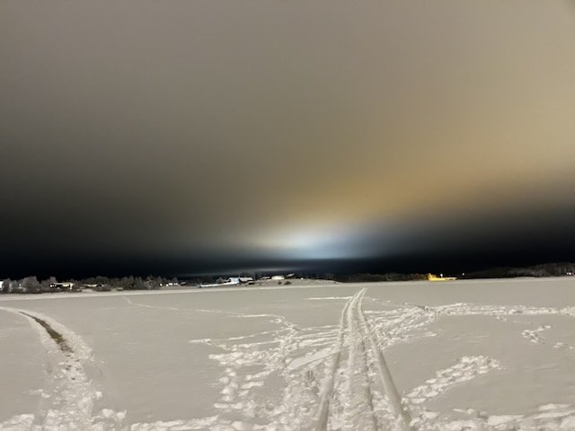 Snow tracks leading across a frozen lake at night toward glowing lights under an overcast winter sky.
