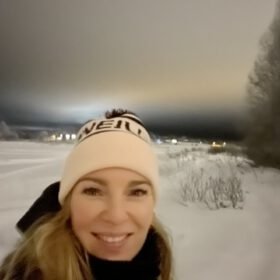 Smiling blonde woman wearing a winter beanie taking a selfie outdoors in a snowy landscape at night.