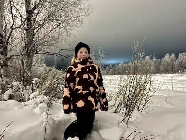 Young girl in a patterned winter jacket standing outdoors in deep snow surrounded by frosty trees.