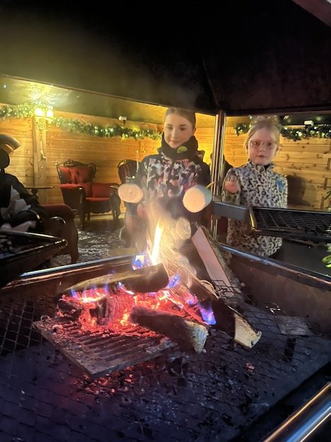 Two young girls roasting marshmallows over an indoor fire pit inside a cozy cabin.