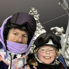 Father and young daughter smiling on a ski chairlift during a night skiing trip in winter gear with helmets and goggles.