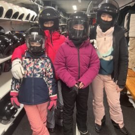Four people wearing winter coats and full-face helmets posing in a snowmobile gear rental shop.