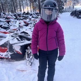 Person wearing a purple jacket and full-face helmet standing in front of snowmobiles in a snowy forest.
