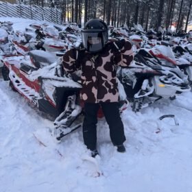Child wearing a helmet and winter coat posing in front of snow-covered snowmobiles in a snowy forest.