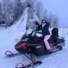 Woman on a black Lynx snowmobile in a snowy winter landscape near a traditional lavvu teepee tent.