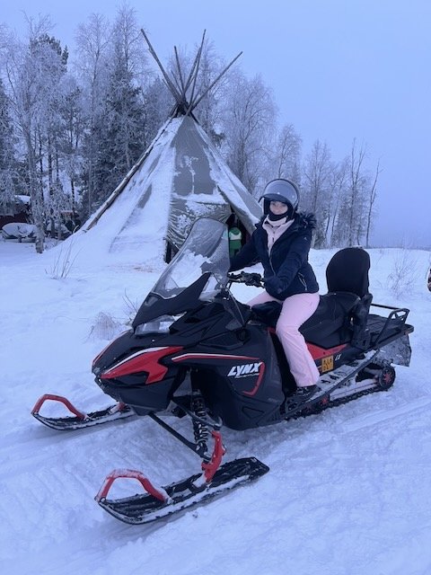 Woman on a black Lynx snowmobile in a snowy winter landscape near a traditional lavvu teepee tent.