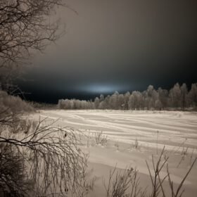 Frozen snow-covered river landscape at night with frosted trees and winter tracks under a dark sky.