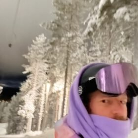 Snowboarder in a purple hoodie and reflective ski goggles on an illuminated snowy night slope near pine trees.