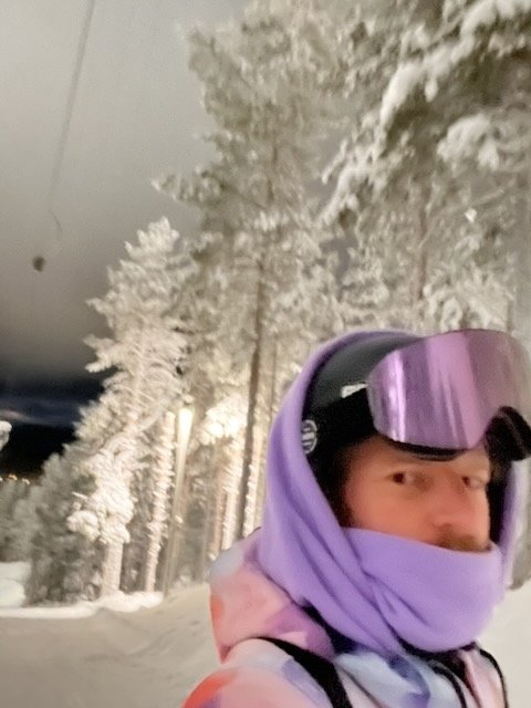 Snowboarder in a purple hoodie and reflective ski goggles on an illuminated snowy night slope near pine trees.