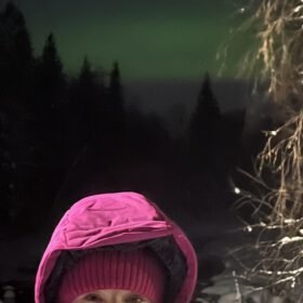 Person wearing a pink winter coat with green northern lights glowing in the dark sky over a snowy pine forest.