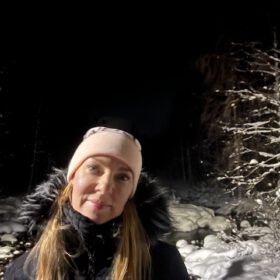Woman wearing a pink beanie and fur-trimmed winter coat standing outside in a snowy forest at night.