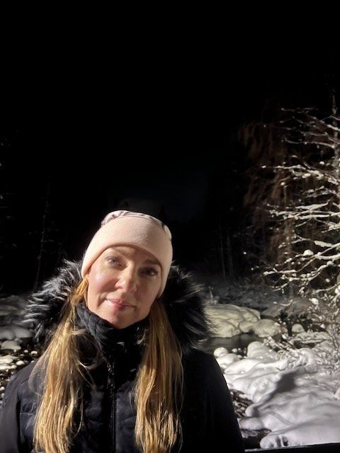 Woman wearing a pink beanie and fur-trimmed winter coat standing outside in a snowy forest at night.