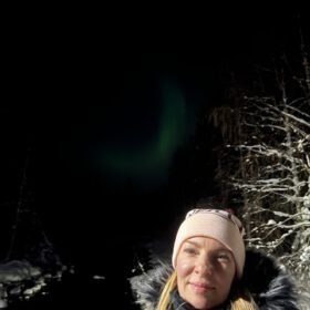 Woman in a pink beanie and winter coat taking a selfie outdoors at night under the northern lights.