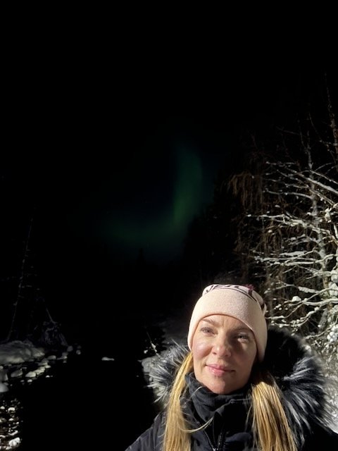 Woman in a pink beanie and winter coat taking a selfie outdoors at night under the northern lights.