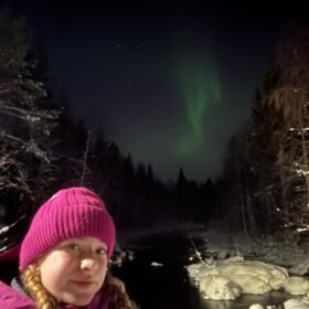 A woman in a pink beanie taking a selfie at night with green Northern Lights glowing over a snowy forest and river.