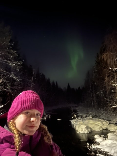 A woman in a pink beanie taking a selfie at night with green Northern Lights glowing over a snowy forest and river.