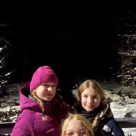 Three young girls wearing warm winter jackets posing outdoors at night in a snowy landscape.