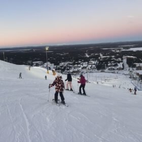 Skiers descending a snowy ski resort slope at dusk with an illuminated mountain village below at sunset.