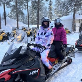 Two people in winter gear and helmets sitting on a Lynx snowmobile in a snowy forest.
